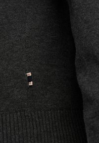 Mørk grå strikkegenser med ribbestrikkede mansjetter og en liten merke med røde og blå detaljer på siden. Myk tekstur, nærbilde.