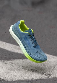 Αθλητικό παπούτσι σε μπλε και νέον πράσινο χρώμα με πλεκτό επάνω μέρος, να αιωρείται πάνω από ασφάλτινη επιφάνεια με μικρά υπολείμματα από κάτω.