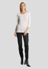 Weißes Langarmoberteil mit rundem Halsausschnitt, kombiniert mit schwarzen Slim-Fit-Hosen und schwarzen, stiefelartigen High Heels, vor einem grauen Hintergrund.
