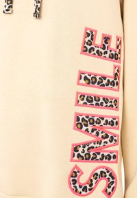 Sudadera beige con texto en negrita bordeado de rosa y un estampado de leopardo en la letra. Los cordones de la capucha son de color blanco.