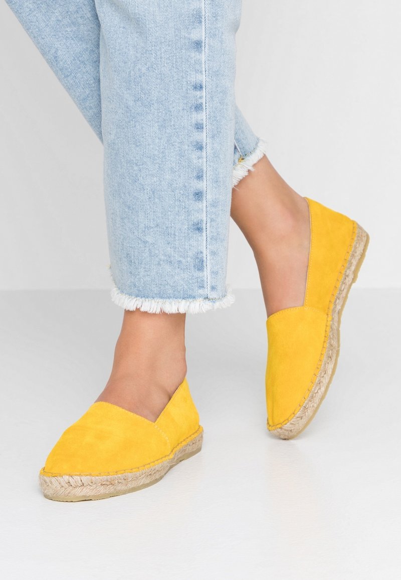 Espadrilles en daim jaune avec un bout en forme de V, semelle en corde de jute et texture lisse. Associées à un pantalon en denim clair raccourci.