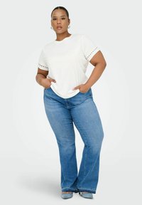 Camiseta blanca de manga corta con acentos perforados en las mangas, combinada con jeans acampanados azules. Mostrada con las manos en los bolsillos.