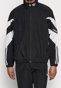 Man i svart Adidas-träningsdräkt med vita ränder på ärmarna och logotyp på bröstet och armen, står mot en neutral bakgrund.