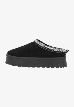 Zapato negro de ante tipo slip-on con suela de goma gruesa, que presenta costuras decorativas blancas alrededor del cuello y una lengüeta en la parte trasera.