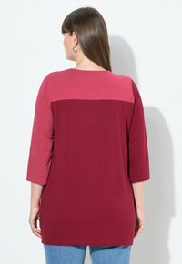 Burgunderfarbene Langarmbluse mit einem helleren rosa horizontalen Streifen über dem oberen Rücken, glatte Textur und lässige Passform, kombiniert mit blauen Jeans.