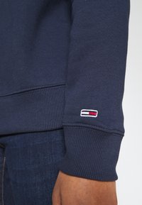 Mörkblå sweatshirt med ribbade muddar och nederkant, med en liten broderad logotyp i röd, vit och blå på ärmen. Mjuk textur.