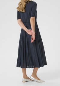 Robe plissée bleue marine avec un corsage ajusté, des manches courtes et une taille élastique. Associée à des ballerines argentées. Tissu lisse et léger.