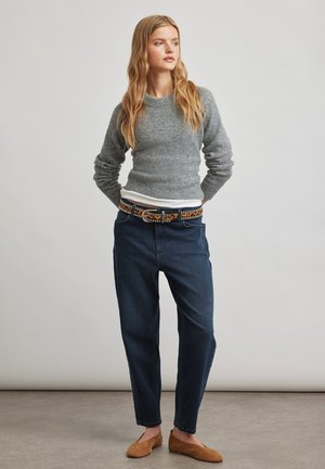 Junge Frau steht mit den Händen hinter dem Rücken, trägt einen grauen Pullover, dunkelblaue Jeans, einen gemusterten Gürtel und braune Ballerinas vor einfachem Hintergrund.