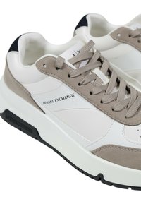 Des baskets blanches et beiges en cuir lisse et en daim, avec des lacets proéminents, une impression de logo et des détails texturés sur le dessus.