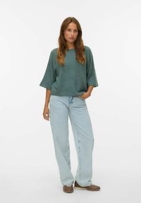 Losse, teal kleurige trui met brede mouwen, gecombineerd met lichtgewassen, rechte jeans. Eenvoudige schoenen maken de outfit compleet.