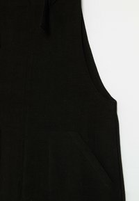 Vêtement noir sans manches avec une grande poche avant et une emmanchure incurvée sur fond blanc.