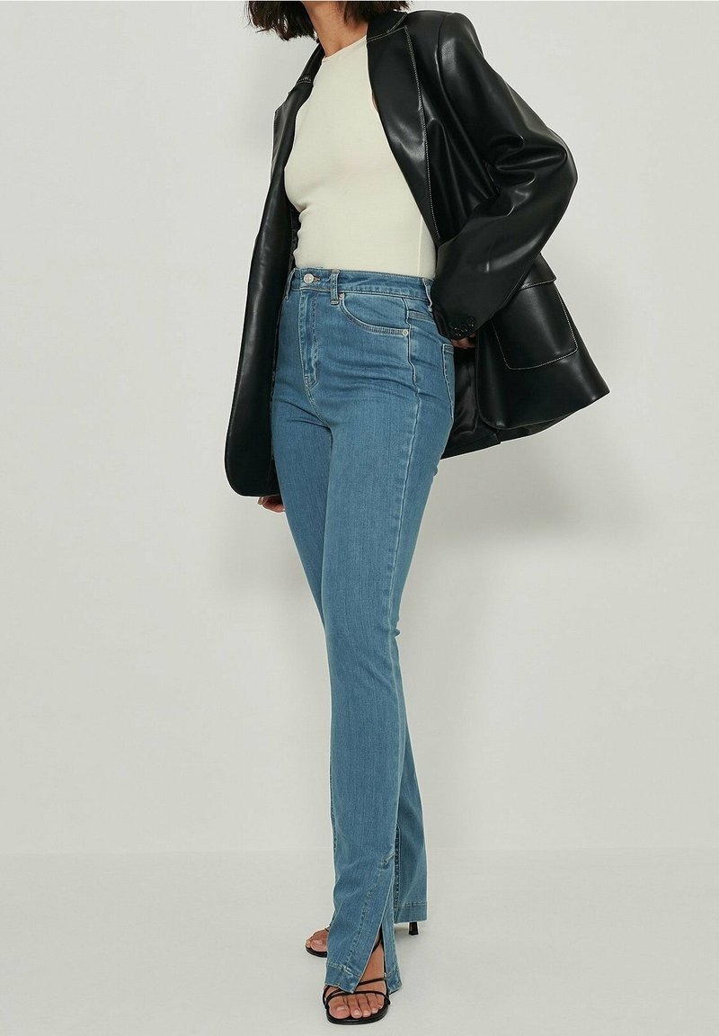 Femme portant une veste en cuir noire, un haut blanc, un jean bleu taille haute fendu au bas et des sandales à talons noires, debout contre un mur neutre.