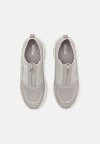 Pár světle šedých slip-on tenisek s síťovou špičkou, přední zip s gravírovaným nápisem "MK" a značkovými stélkami na bílém pozadí.