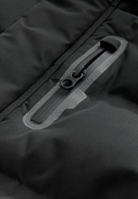 Gros plan sur la poche d'une veste imperméable noire avec une fermeture éclair scellée et une tirette en tissu, cousue pour la durabilité et la résistance à l'eau.