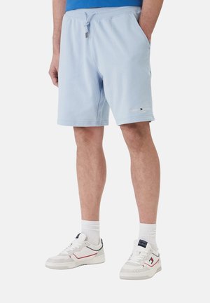 Hombre con pantalones cortos azul claro con cordón, calcetines blancos y zapatillas blancas con detalles en rojo y azul marino, de pie con las manos en los bolsillos.