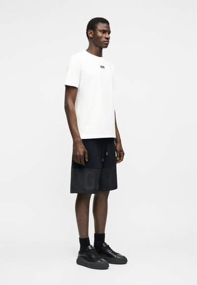 Άνδρας στέκεται στραμμένος προς τα δεξιά φορώντας λευκό t-shirt με μικρό μαύρο λογότυπο, μαύρα σορτσάκια, μαύρες κάλτσες και μαύρα αθλητικά παπούτσια, σε λευκό φόντο.