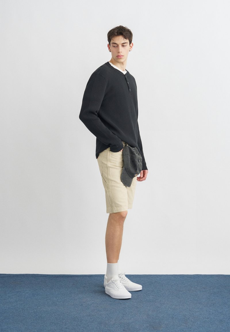 Jonge man staand, gekleed in een zwart shirt met lange mouwen, beige korte broek, witte sokken en sneakers, houdt een grijze pet vast en kijkt opzij.