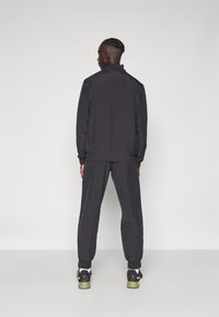 Schwarze Trainingsjacke und passende Hose aus leichtem Stoff, mit elastischen Bündchen und hohem Kragen, kombiniert mit schwarzen und grünen Turnschuhen.