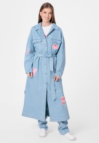 Manteau en denim bleu clair avec une ceinture, présentant des patches décoratifs roses. Il a un col, une fermeture par boutons et des poches latérales.