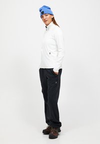 Giacca bianca con zip, pantaloni neri con tasche laterali, berretto blu e stivali da trekking marroni. Il tessuto appare liscio con un design aderente.