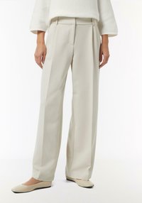 Weite Hose aus hellem beigen Stoff, mit hoher Taille, Falten und glatter Textur. Kombiniert mit passenden geschlossenen Ballerinas.