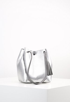 Silberne Metallic-Eimer-Tasche mit einem einzelnen Riemen und einem Quasten-Detail, auf einem weißen Podest vor einem schlichten weißen Hintergrund platziert.