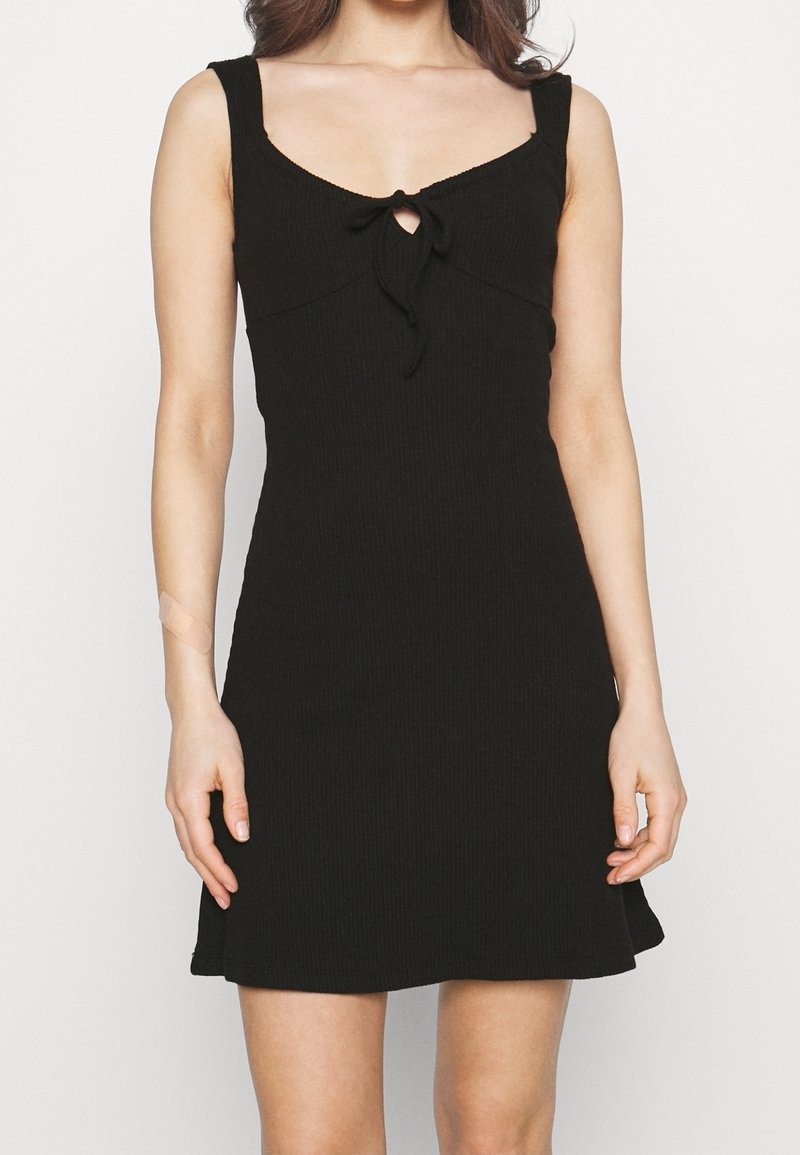 Femme portant une robe mini noire sans manches à côtes avec un détail de nœud à l'avant et une petite découpe en forme de clé sous le col.