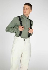 Olijfgroene shirt met lange mouwen en een ritssluiting aan de kraag, gecombineerd met witte broeken vastgemaakt met zwarte bretels. Kenmerkt zich door soepele texturen en contrasterende kleuren.