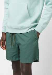 Felpa verde chiaro con tasca frontale, abbinata a pantaloni corti verdi scuri con chiusura a cordoncino e tasche laterali. Tessuto dalla texture liscia.