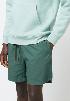 Felpa verde chiaro con tasca frontale, abbinata a pantaloni corti verdi scuri con chiusura a cordoncino e tasche laterali. Tessuto dalla texture liscia.