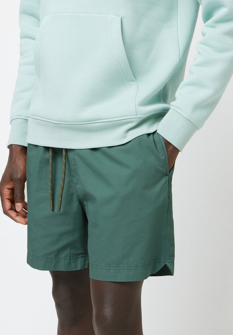 Felpa verde chiaro con tasca frontale, abbinata a pantaloni corti verdi scuri con chiusura a cordoncino e tasche laterali. Tessuto dalla texture liscia.