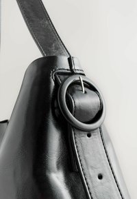 Close-up of a black leather bag strap with a round black buckle and visible stitching on the smooth, shiny surface.
