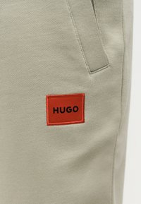 Λεπτομέρεια μπεζ υφασμάτινου παντελονιού με μπροστινή τσέπη και κόκκινο ορθογώνιο ταμπελάκι με την ένδειξη "HUGO" ραμμένο κοντά στην τσέπη.