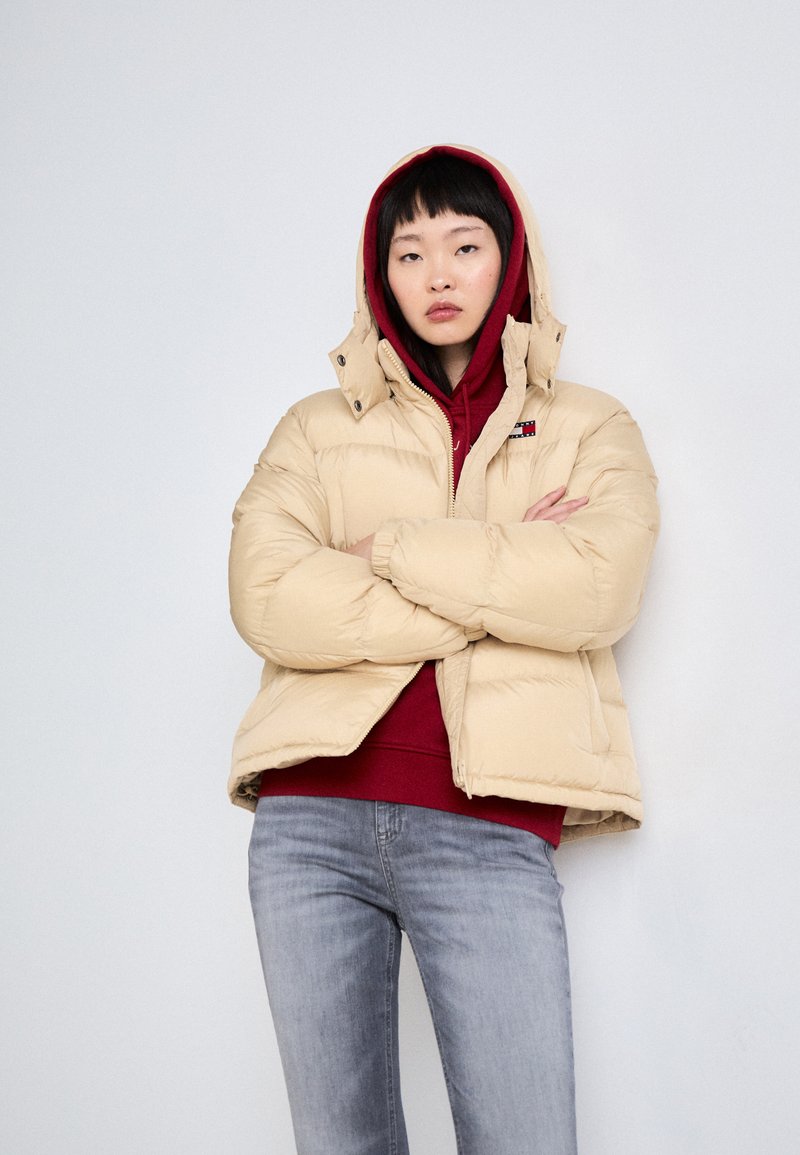 Beige Daunenjacke mit hohem Kragen und Kapuze, mit rotem Innenfutter und einem Logo-Patch, getragen über einem roten Sweatshirt und grauen Jeans.