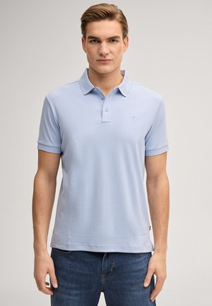 Młody mężczyzna stoi twarzą do przodu, ubrany w jasnoniebieską koszulkę polo i niebieskie dżinsy na jednolitym jasnym tle.