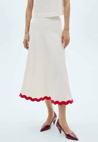 Falda de punto blanca con un dobladillo rojo en forma de onda, que cae por debajo de la rodilla. Combinada con tacones color burdeos, que presentan una punta afilada y un diseño de tacón curvado.