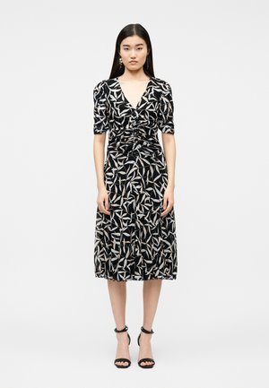 Γυναίκα που στέκεται, φορώντας μαύρο φόρεμα μίντι με λευκά και μπεζ μοτίβα φύλλων, με ρυτιδωμένο μπροστινό μέρος, φουσκωτά μανίκια και μαύρα πέδιλα με λουράκι στον αστράγαλο.