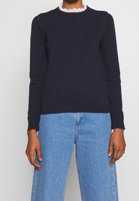 Mörkblå tröja med långa ärmar, ribbade manschetter och nederkant, prydd med en vit scalloped spetskrage. Matchas med ljusblå vida jeans.