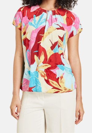 Femme portant un chemisier à manches courtes avec un grand imprimé floral aux couleurs rouge vif, rose, bleu, jaune et beige, associé à un pantalon blanc cassé.