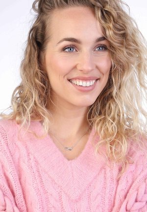 Roze gebreide trui met een V-hals, voorzien van een textuurpatroon. Een zilveren oneindigheidssieraad rust dicht bij de hals. Zacht, golvend haar.