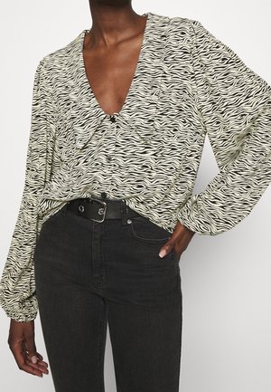 Person iført en langærmet zebra-print bluse, der er stukket ned i højtaljede sorte jeans med et sort bælte, hånd hvilende på hoften.