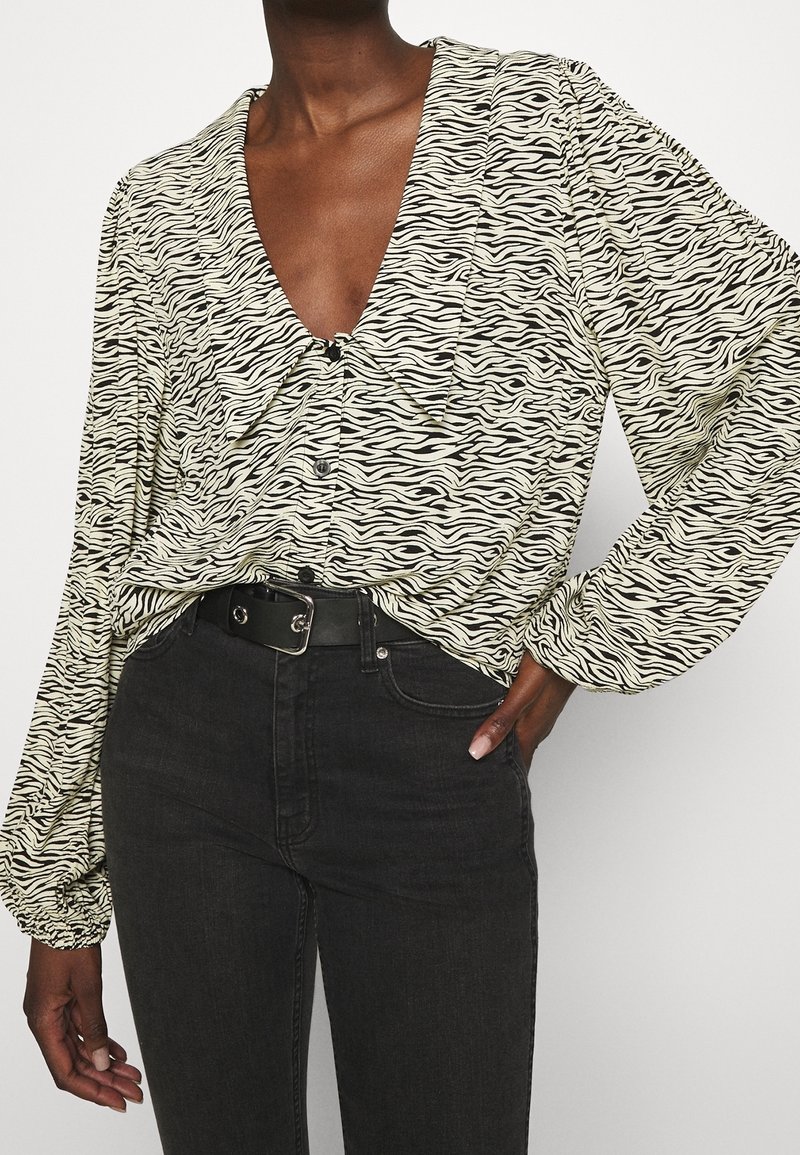 Personne portant un chemisier à manches longues à imprimé zèbre, rentré dans un jean noir taille haute avec une ceinture noire, main posée sur la hanche.