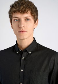 Zwarte button-up shirt gemaakt van textuurstof, met een puntige kraag, borstzak en zilverkleurige knopen. Effen achtergrond.