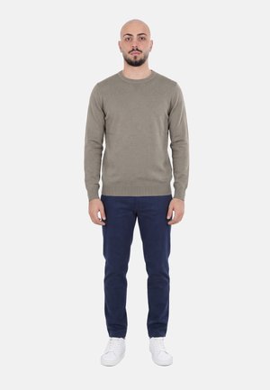 Uomo con testa rasata e barba che indossa un maglione verde oliva, pantaloni blu navy e scarpe da ginnastica bianche, in piedi davanti a uno sfondo semplice.