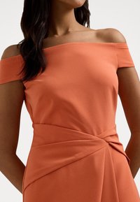 Avskuren axel, korallklänning i mjukt tyg med en tvistad detalj vid midjan, som har en slät struktur och en kroppsnära siluett.