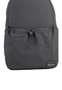 Sac à dos gris foncé en tissu léger et texturé. Il dispose d'une fermeture à cordon et d'une petite étiquette rectangulaire de la marque en bas.