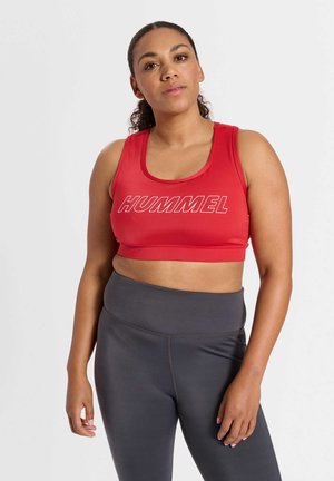 Vrouw met krullend haar die een rood cropped sporttopje met "HUMMEL"-logo en hooggetailleerde donkergrijze leggings draagt, staand tegen een witte achtergrond.