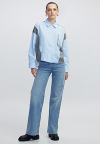 Camisa de botones azul claro con paneles laterales grises y una cadena decorativa en el bolsillo, combinada con jeans de pierna ancha azul claro y botas negras.