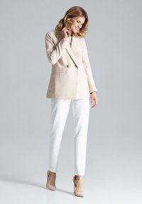 Beige dubbelgebreide blazer met een gladde afwerking, gecombineerd met strakke witte broeken en nude hoge hakken, tegen een lichtgrijze achtergrond.