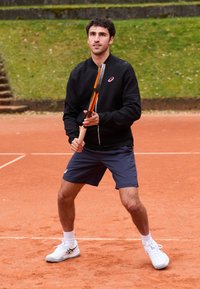 Črna zip-up jakna, mornarsko modre kratke hlače, drži teniško lopar s oranžnimi poudarki; stoji na zemeljskem igrišču z zelenico v ozadju.